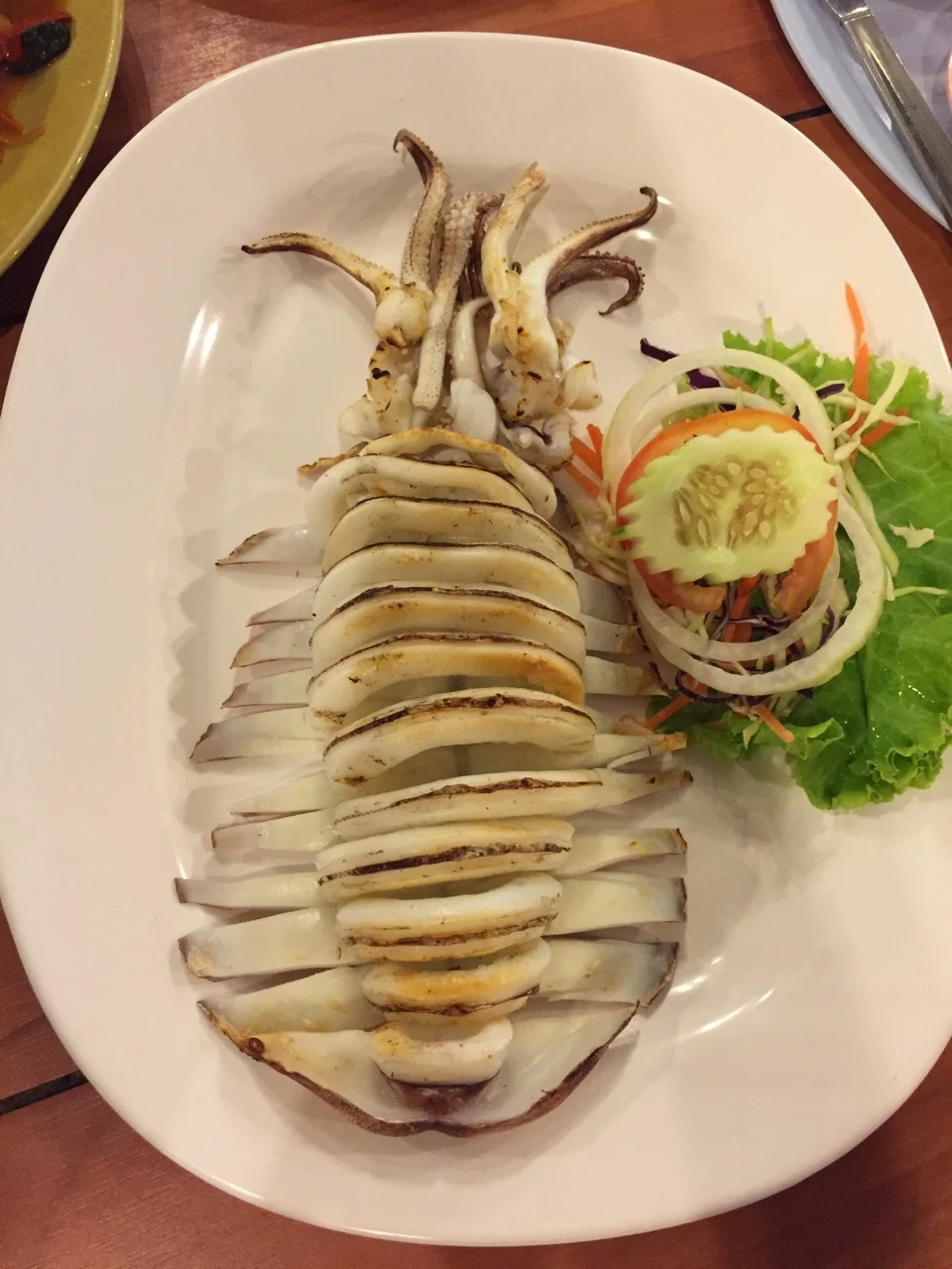 A whole grilled squid on a white plate with a small side salad, photographed from above on a wooden table.