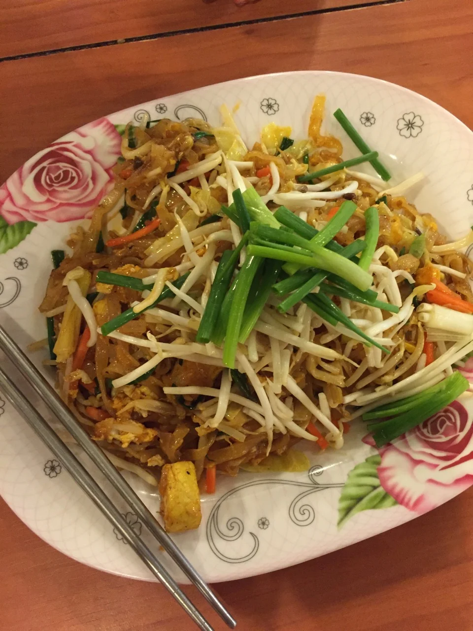 Pad thai on a floral plate with metal chopsticks, shot from above on a wooden table.