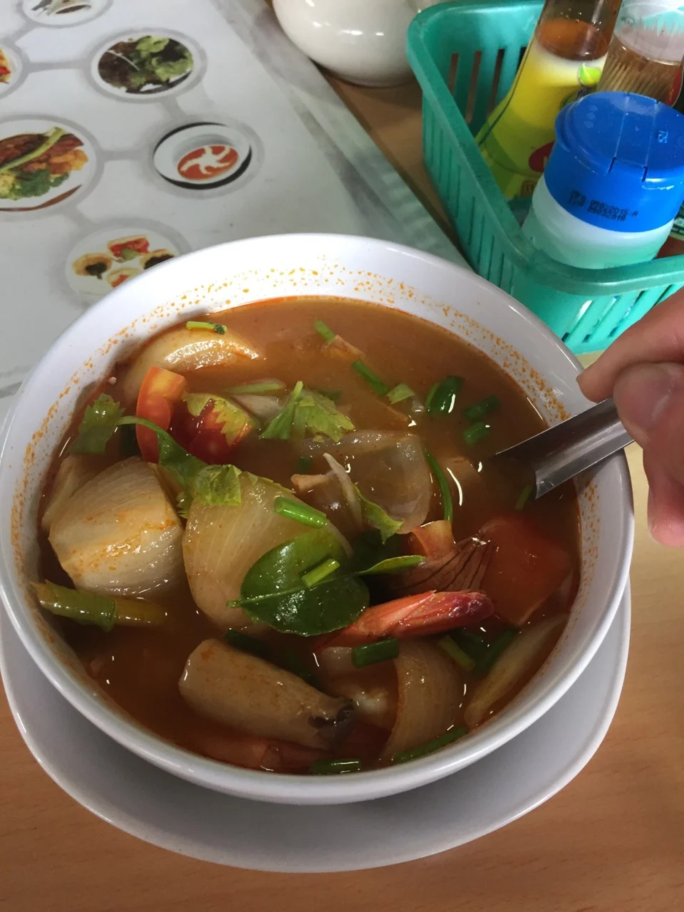 A bowl of tom yum soup with a hand holding the spoon, condiment bottles, and a printed placemat in the background.